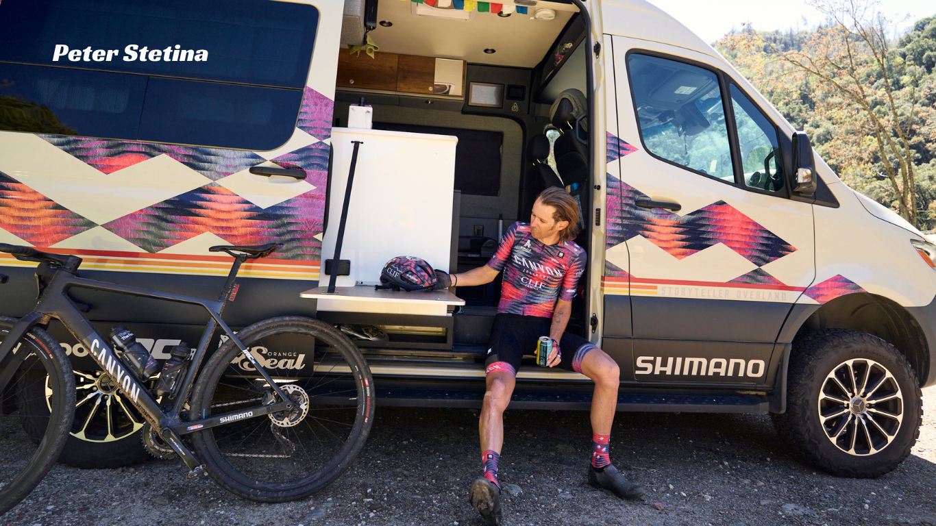 A cyclist in a pink and black jersey sits on the open side door of a white camper van with colorful geometric graphics, next to a black mountain bike. The van has 'Peter Stetina' on the window and 'SHIMANO' on the door, with an outdoor wooded background.