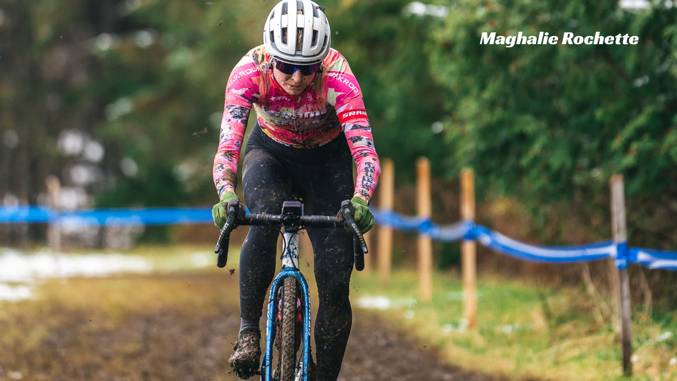 A cyclist wearing a pink and black patterned jersey, black pants, and a white helmet rides a blue bicycle on a muddy trail during an outdoor cycling event. The text 'Maghalie Rochette' is visible in the top right corner.
