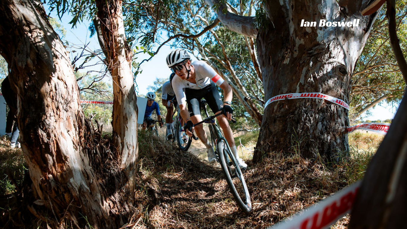 A cyclist wearing a white jersey and helmet rides a mountain bike through a wooded trail, with large trees framing the scene and red-and-white race tape visible. The text 'Ian Boswell' appears in the upper right.