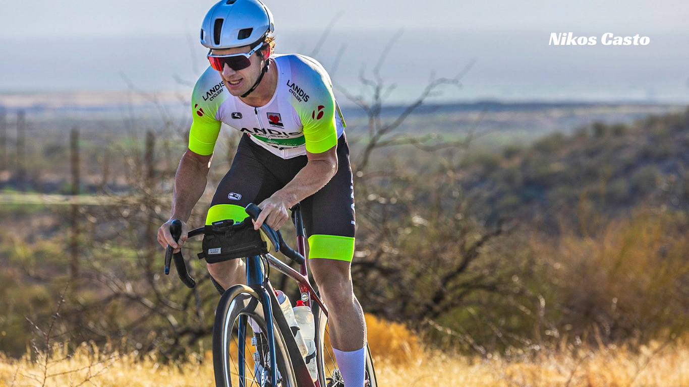 A cyclist wearing a white, black, and neon green cycling jersey and shorts rides a road bike on a dry, grassy hillside. The rider is wearing a light blue helmet and sunglasses. Nikos Casto